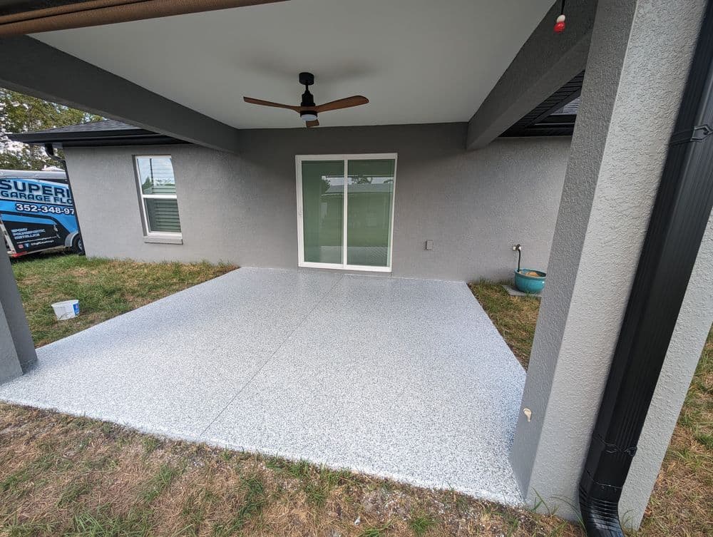Patio with gray epoxy flooring, ceiling fan, and sliding doors, surrounded by grass.